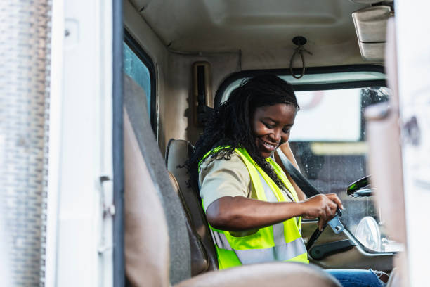 A mature black woman, in her 40s, working as a truck driver at a lumberyard sitting in the driver's seat of a delivery truck, fastening her seat belt