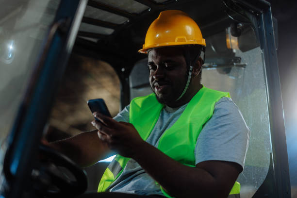 Young African-American forklift driver sitting in the forklift and typing a message on his cell phone.